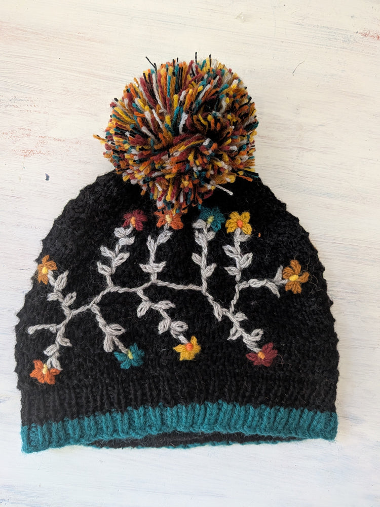 Black knit beanie with floral patterns and a colorful pom-pom on a light wooden background