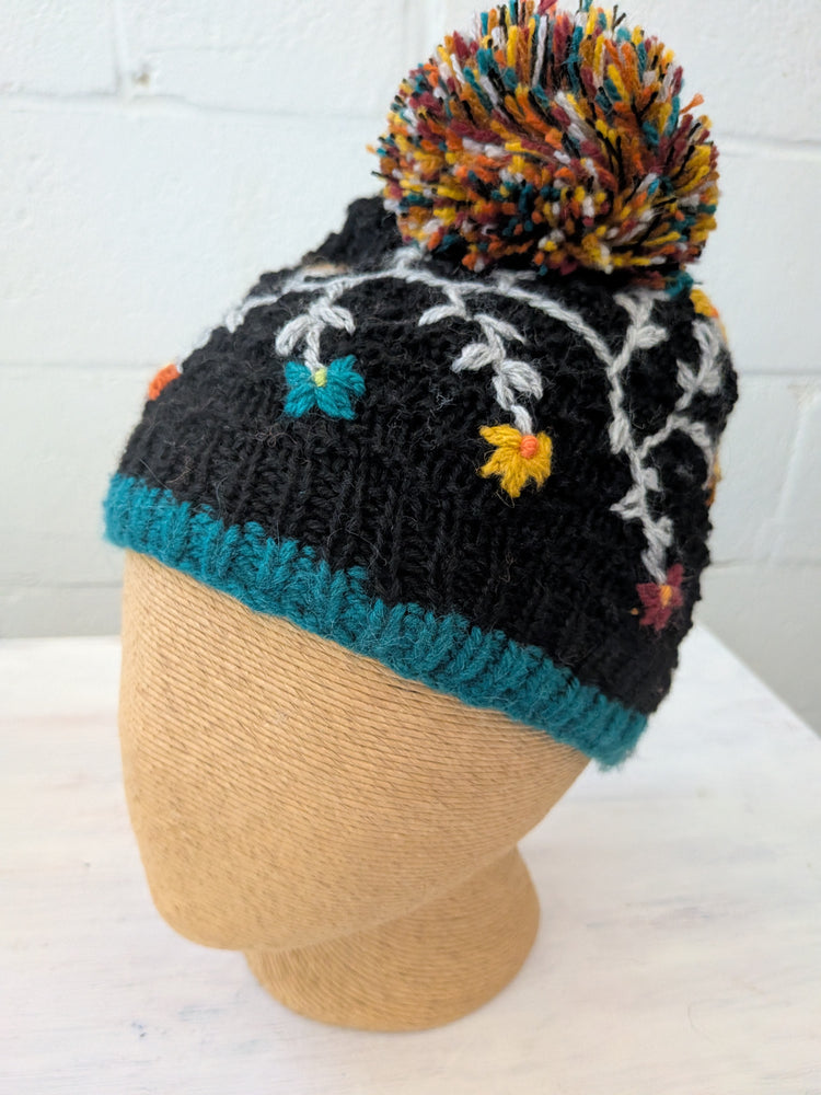 Black knit beanie with colorful pom-pom and floral embroidery on a mannequin head against a white tiled wall.