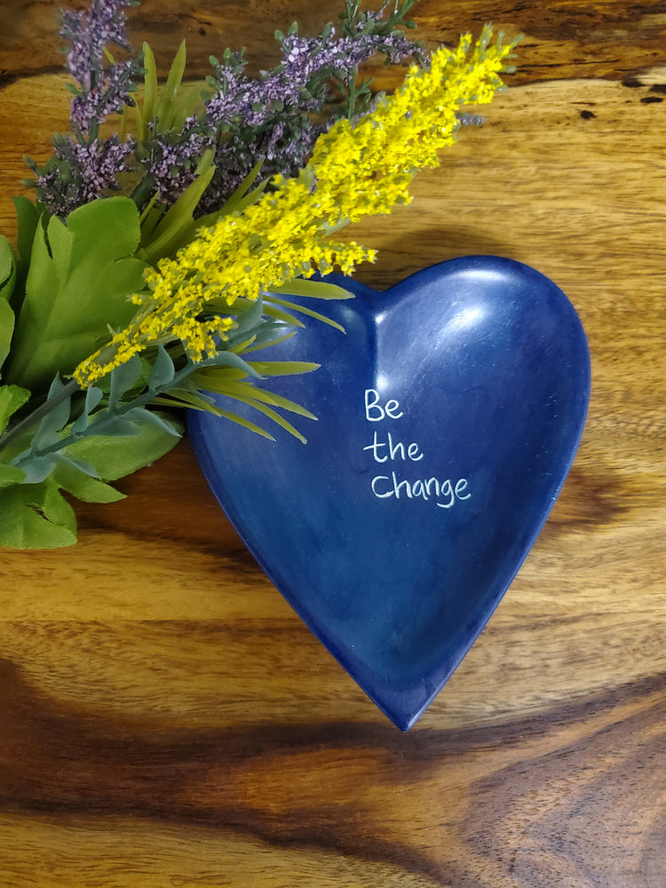 A blue heart-shaped stone dish with the phrase 'Be The Change' engraved on it, placed on a wooden surface with a bundle of flowers beside it.