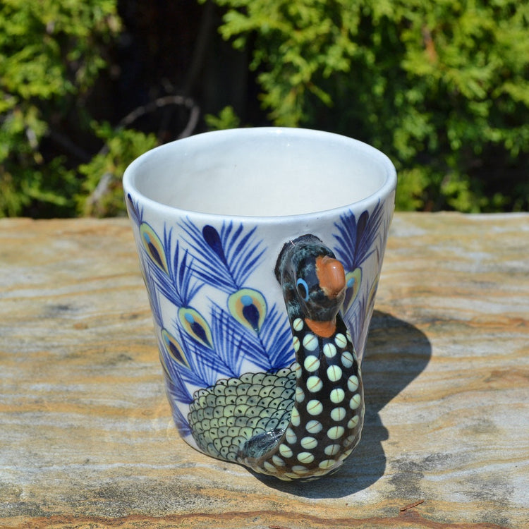 Ceramic mug with peacock design on a wooden surface with greenery in the background