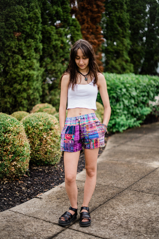 Woman wearing a white crop top and colorful shorts standing on a sidewalk with greenery in the background