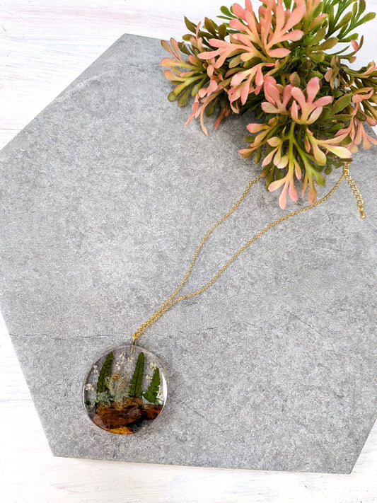 Necklace with a round pendant featuring greenery on a gray hexagonal stone surface.