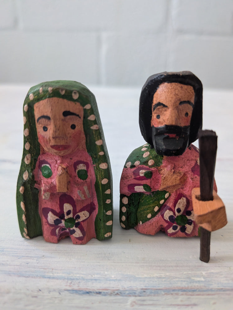 Two wooden nativity figures, one with a green headscarf and pink dress, the other with a beard and holding a staff, on a white background.