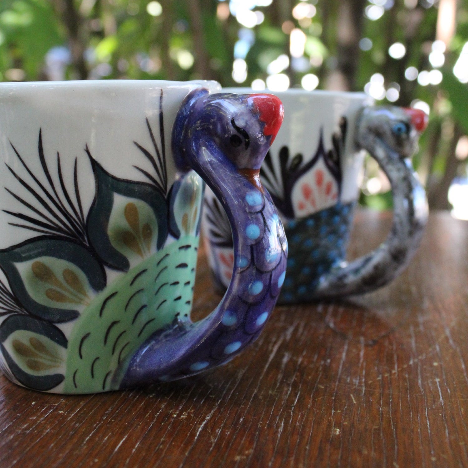 Two ceramic mugs with peacock handles on a wooden surface with a blurred natural background