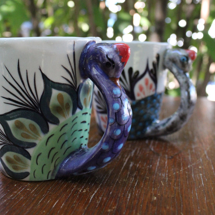Two ceramic mugs with peacock handles on a wooden surface with a blurred natural background