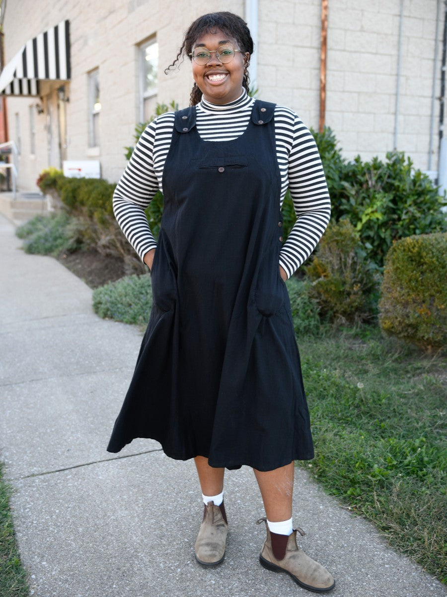Person wearing a black jumper dress over a striped shirt on a sidewalk.