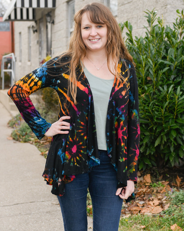 Woman wearing a colorful jacket standing on a sidewalk with a building and greenery in the background