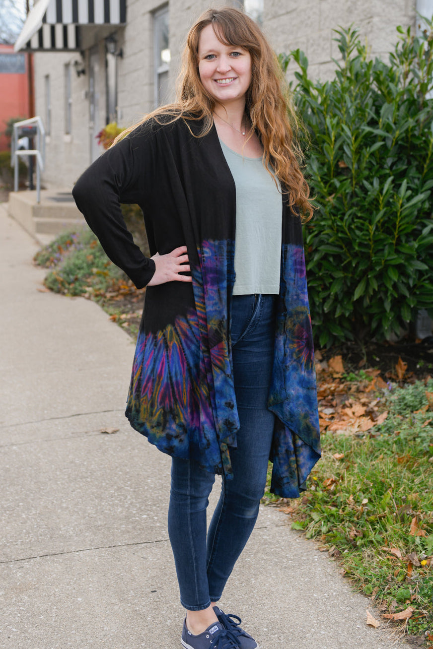 Woman wearing a colorful tie-dye cardigan over a green top and jeans, standing on a sidewalk.