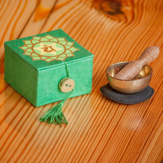 green paper box with chakra icon sitting next to small brass singing bowl with wooden handle and black fabric pillow